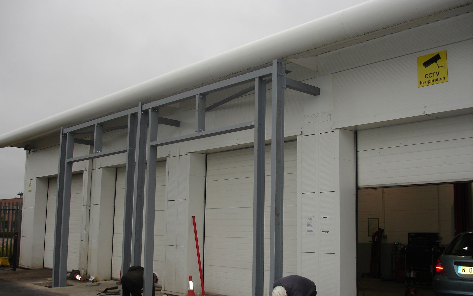 Exterior view of an industrial building with large gray framework structures and a CCTV in operation sign above.