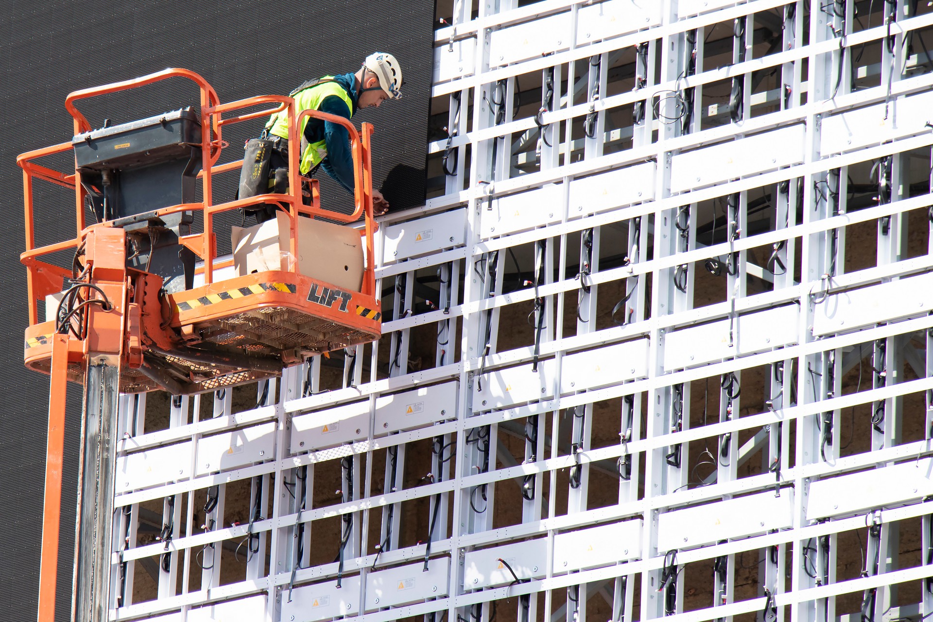 Construction worker in crane basket installing sheets cladding for covering structure on a building facade wall